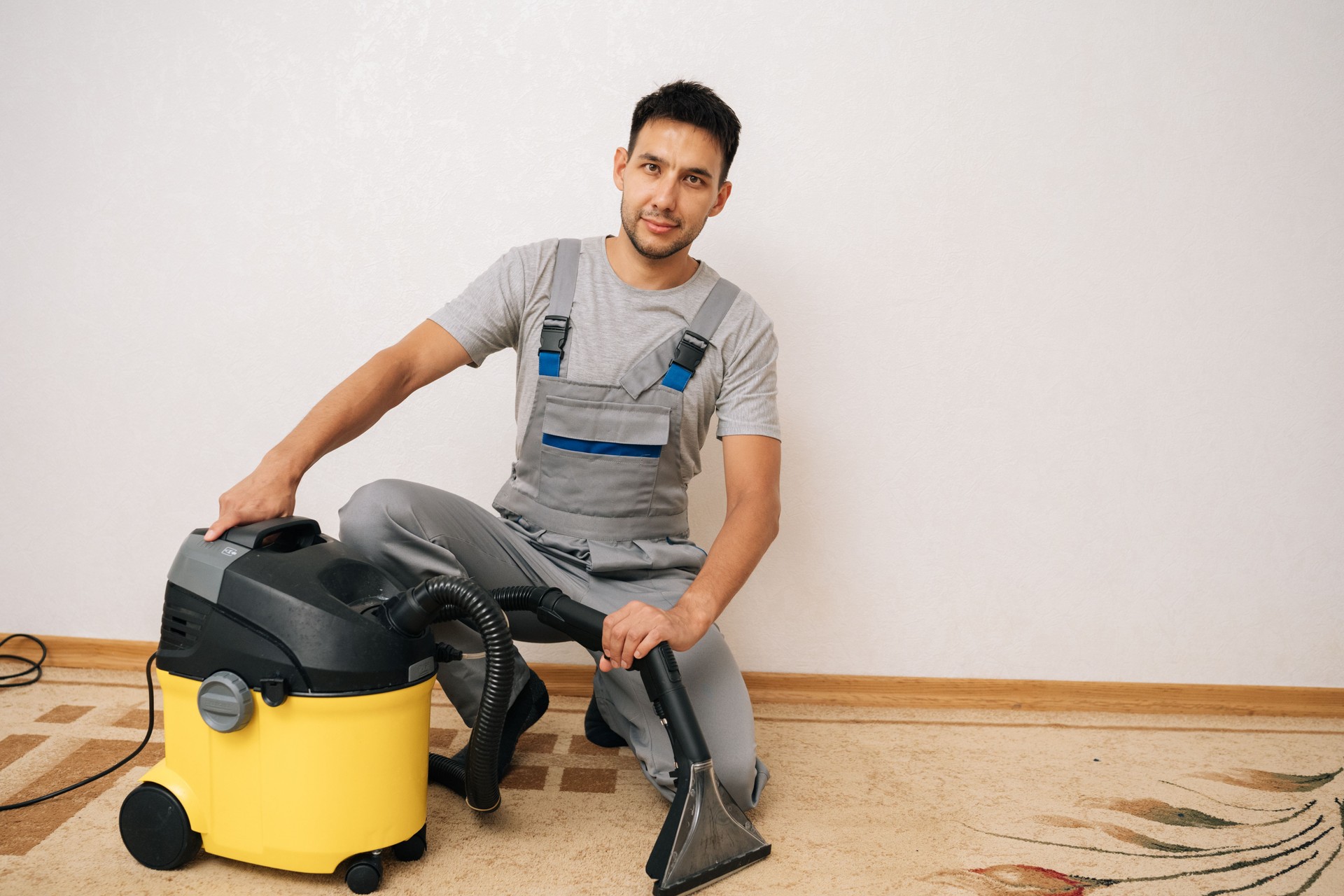 Portrait of professional cleaner male sitting on floor next to washing vacuum cleaner, holding cleaning attachment, prepared to deliver expert carpet cleaning services